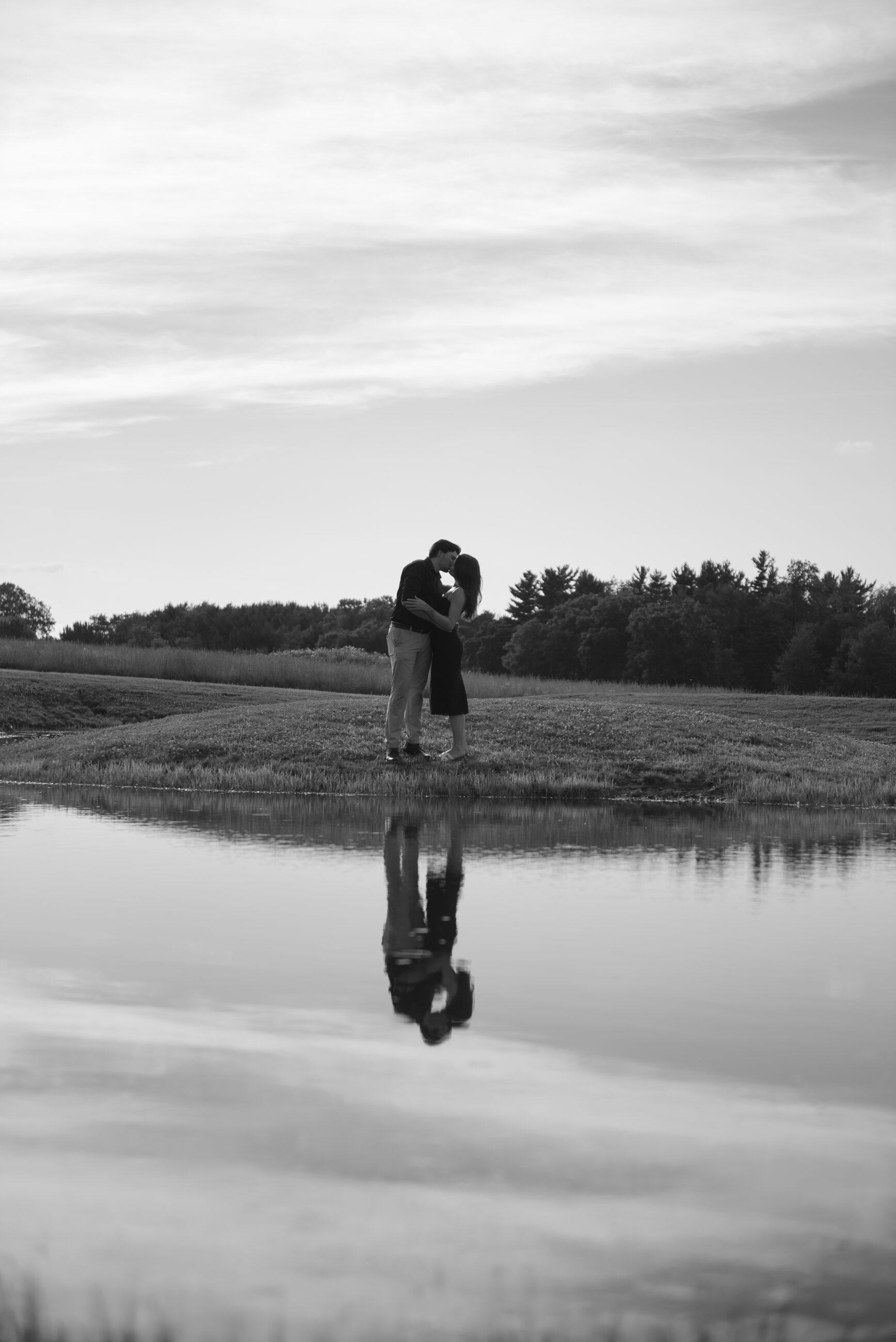 A couple mirrored in still pond water beneath a wide sky, black and white
